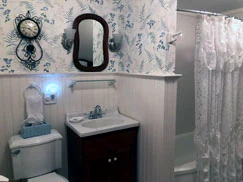A bathroom with blue-and-white botanical wallpaper above white beadboard, an oval mirror in a dark frame, a dark wood vanity with a white top, and a lace shower curtain