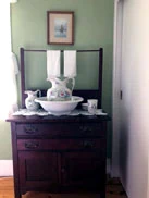 A washstand with a china pitcher and bowl beneath a framed picture