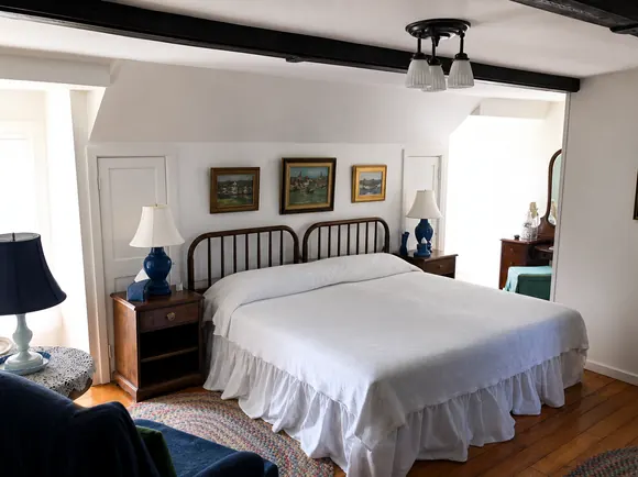 A light guest room under a beamed ceiling with an iron bedstead, a white spread and two windows
