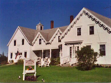 The Mill Pond House from the road: a white c.1860 farmhouse with a cupola, scalloped gingerbread trim along every gable, an American flag by the door and white chairs on the lawn beside the hanging sign