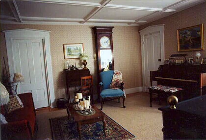 The living room, with a coffered ceiling, a grandfather clock, an upright piano and a blue wing chair on an oriental rug