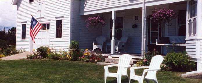 The front porch on a summer afternoon, with hanging fuchsia baskets, white wicker chairs in the shade and two Adirondack chairs out on the grass